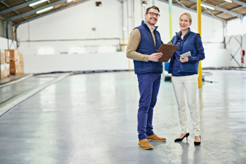 Two warehouse staff standing on a polished concrete floor, holding a clipboard and tablet